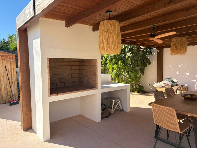 Barbacoa de obra en la Playa de San Juan (Alicante): hogar de ladrillo refractario y encimera blanca bajo porche con vigas de madera